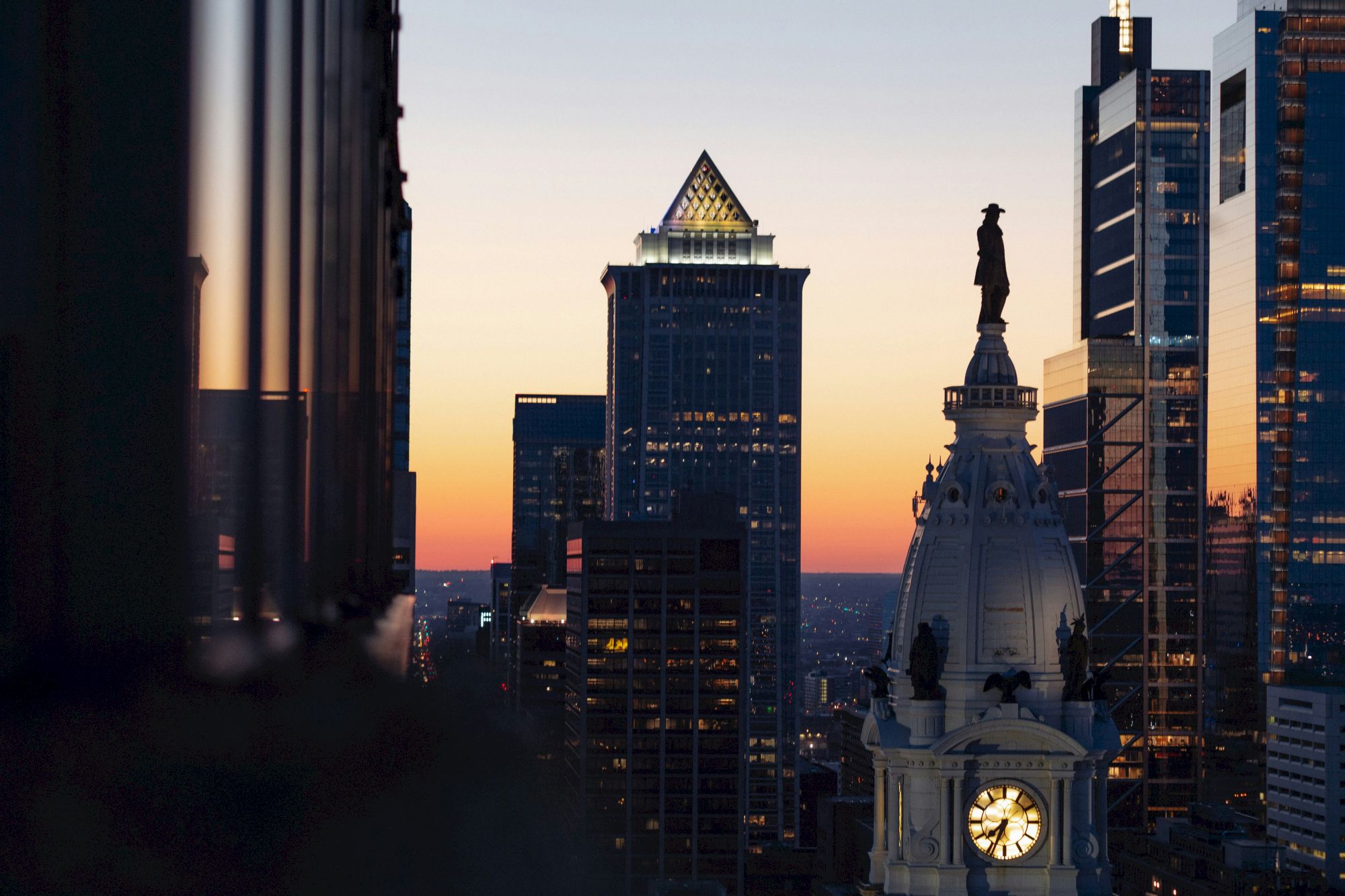 A city skyline at sunset, featuring a clock tower and tall buildings with a vibrant sky in the background.