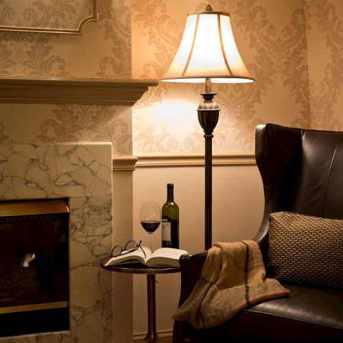 A cozy scene with a leather chair, a lamp, a fireplace, and a side table with wine, a book, and glasses, creating a warm ambiance.