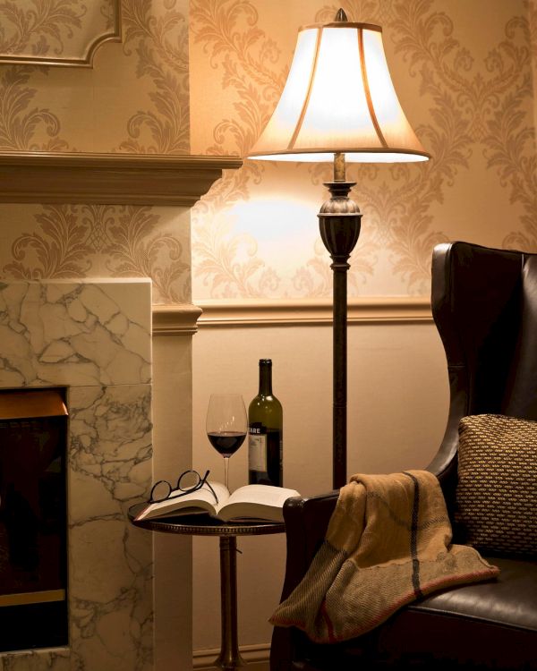 A cozy scene with a leather chair, a lamp, a fireplace, and a side table with wine, a book, and glasses, creating a warm ambiance.