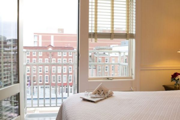 A cozy bedroom with a neatly made bed, a side lamp, flowers, and a view through glass doors showing a building outside.