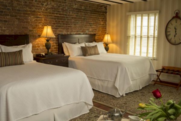 A cozy bedroom has two beds with white linens, a brick accent wall, a large clock, warm lighting, and a tray with flowers.