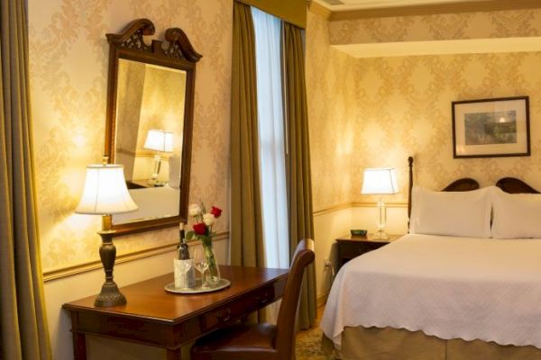 A cozy hotel room featuring a double bed, nightstands with lamps, a mirror, and a table with chairs. The decor is classic and elegant.