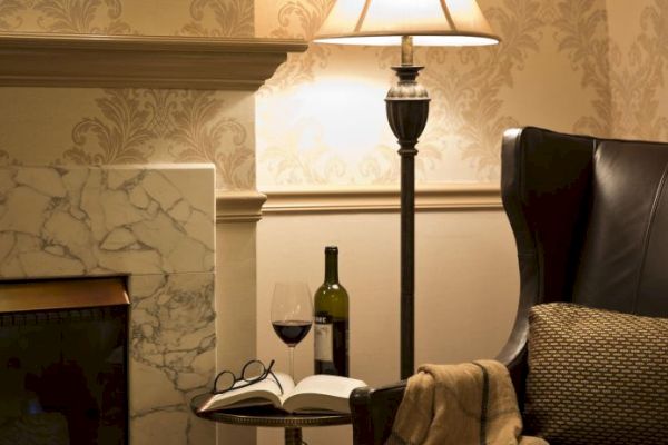 A cozy setting with a leather chair, a lamp, a marble fireplace, a book, glasses, a wine bottle, and a glass on a side table ends the scene.