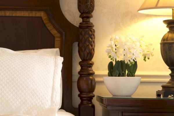 A cozy bedroom scene featuring a wooden headboard, a white pillow, a lamp on a nightstand, and a potted plant with white flowers.