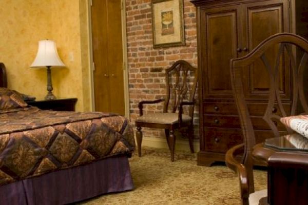 A cozy bedroom with a bed, lamp, wooden furniture, and exposed brick wall. A table holds a laptop, papers, and chair nearby.