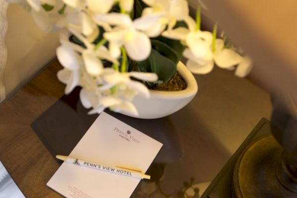 A bedside table with a lamp, notepad, pen labeled "Penn's View Hotel," and a vase of white flowers.