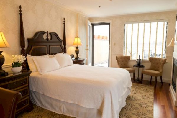 A cozy bedroom with a large bed, elegant lamps, and classic furniture, featuring a window with blinds and a small seating area.