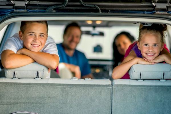 A family is sitting in a car, with two children smiling at the back. The scene is cheerful and relaxed, suggesting a road trip.