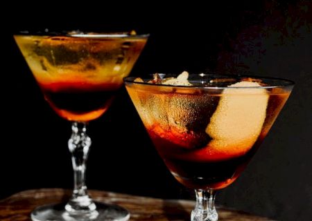 Two cocktails in martini glasses, featuring a dark liquid with ice and orange peel, are set against a dark background.