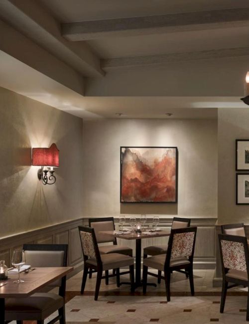 Cozy restaurant interior with neatly set tables, wall art, red-shaded sconces, and a candle-style chandelier, creating a warm ambiance.