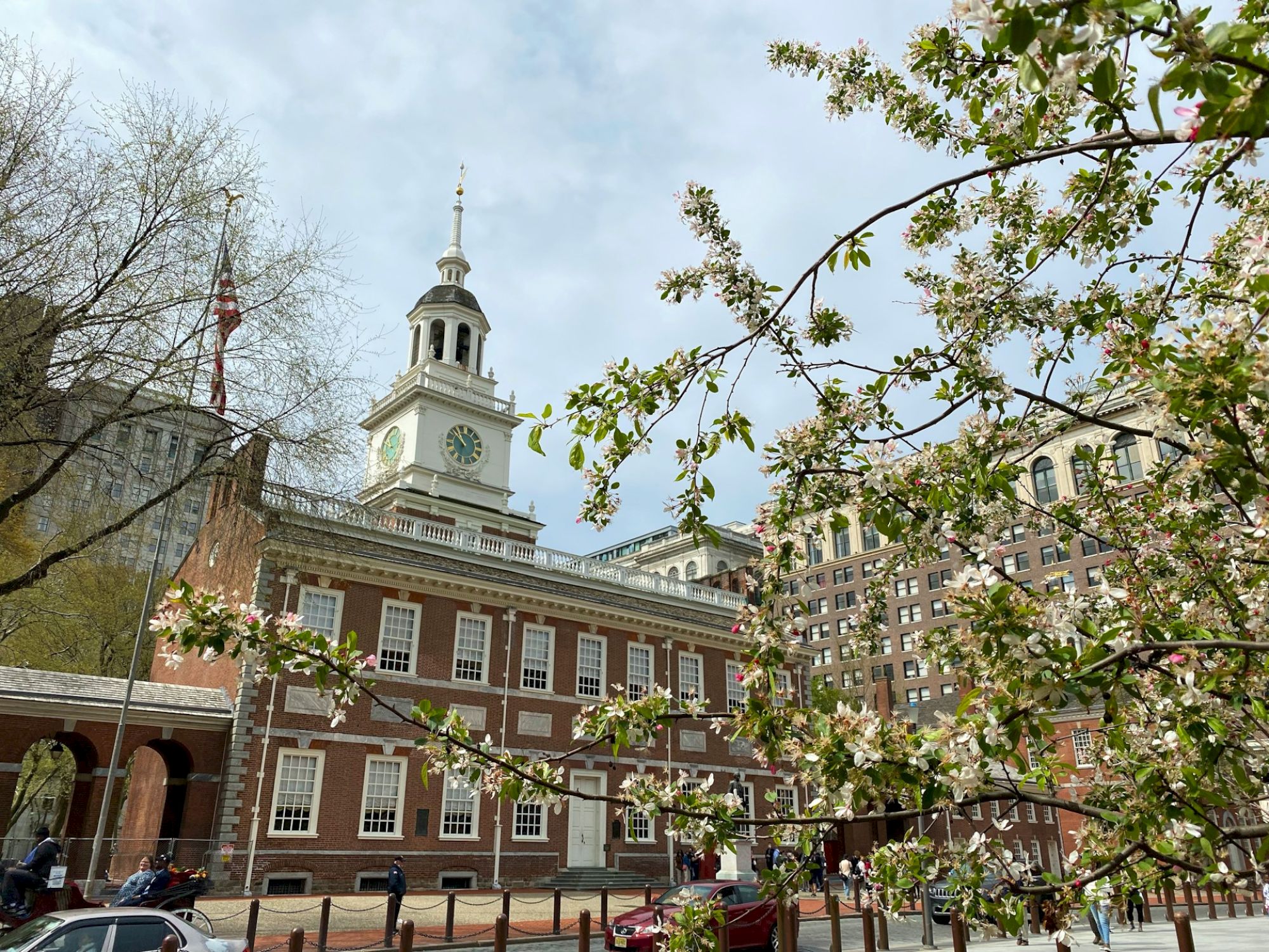 Historic red-brick building with a clock tower, spring blossoms in foreground, and urban backdrop. People are seen near the structure.