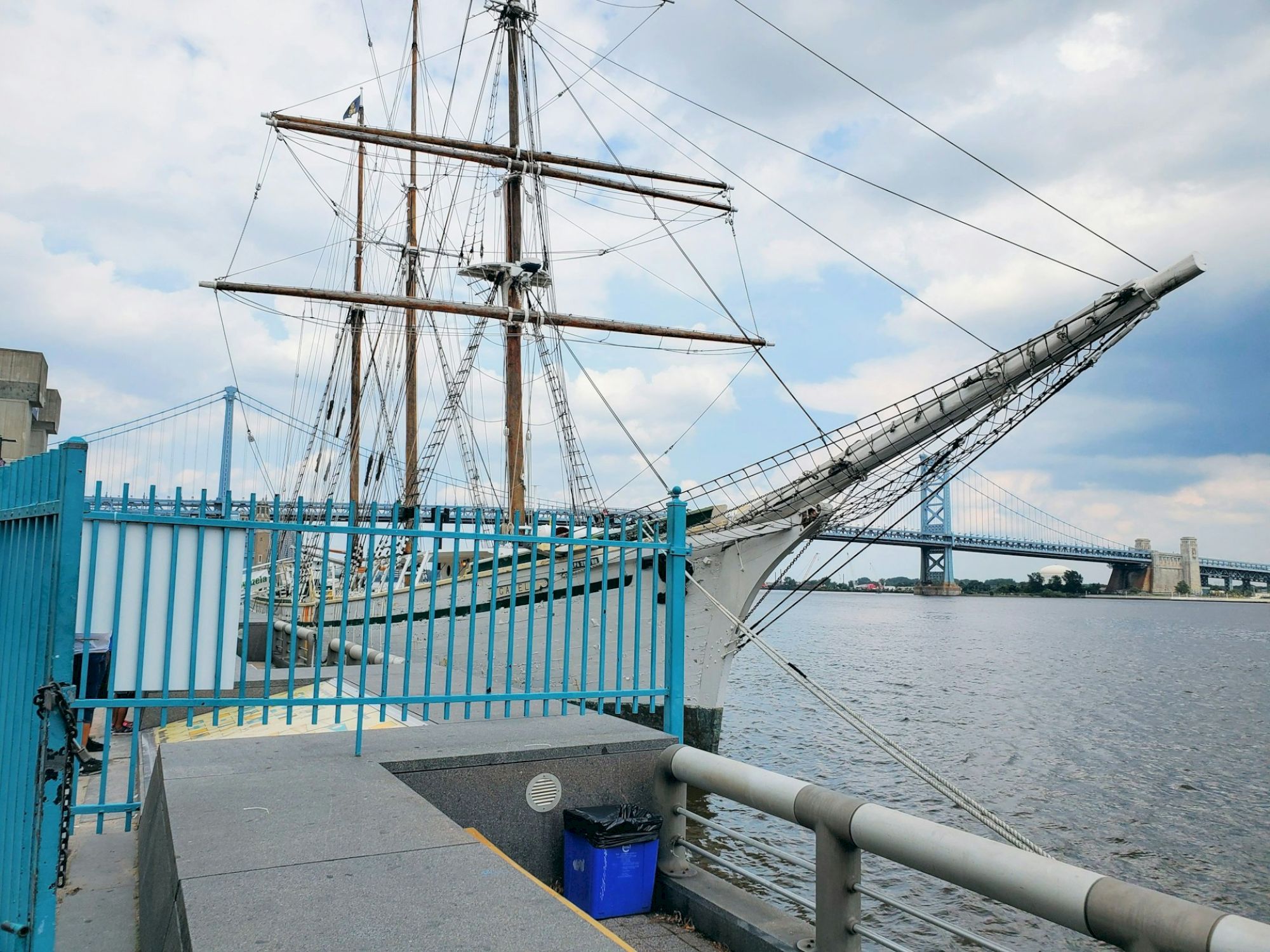 A docked sailing ship with masts is next to a blue fence, and a large bridge spans the water under a cloudy sky.