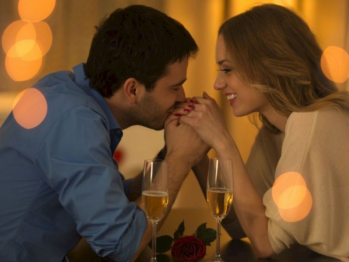 A couple shares a romantic moment at a table with two glasses of champagne and a rose, surrounded by warm lights.