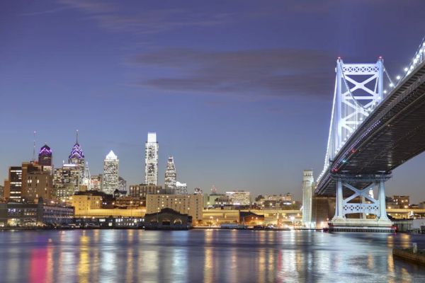 City skyline with illuminated skyscrapers and a lit-up bridge by the waterfront at dusk, reflecting on the water's surface.