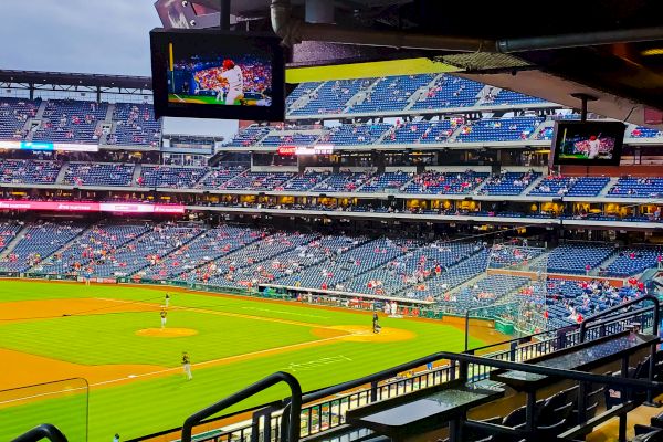 The image shows a baseball stadium interior with players on the field, some spectators, and screens displaying the game.