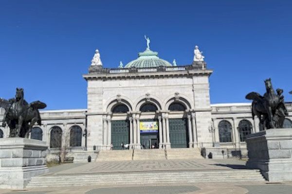 The image shows a grand neoclassical building with a dome, flanked by statues of winged horses on pedestals, under a clear blue sky.