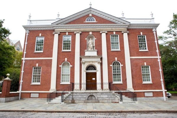 The image shows a historic brick building with white columns and a statue above the entrance, flanked by multiple windows.