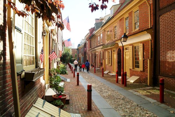 A narrow brick street lined with historic houses, adorned with American flags, and people walking down the path.