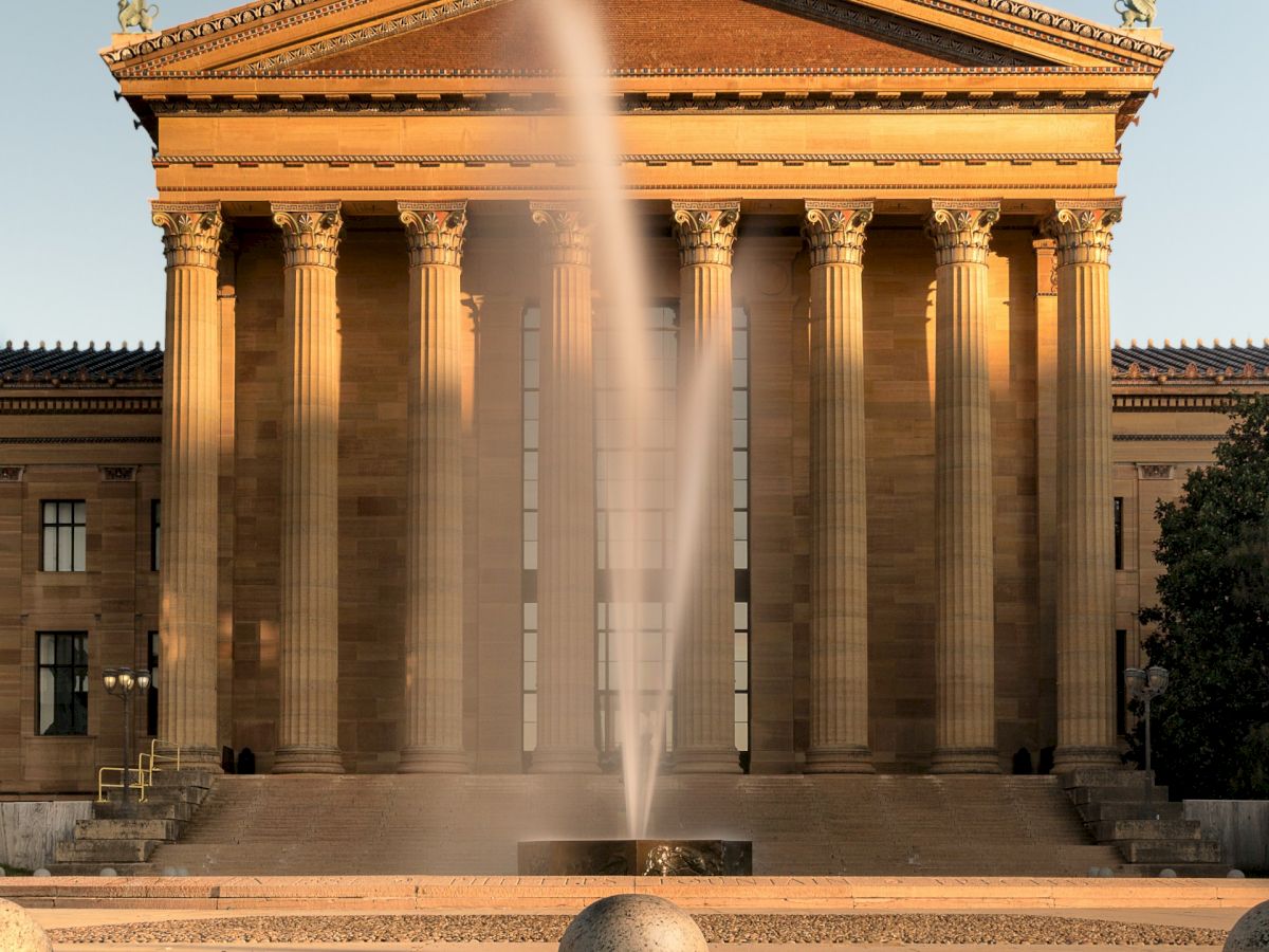 The image shows a neoclassical building with columns and a central fountain in front.